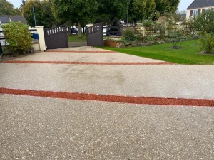 Entrée paysagée en béton désactivé, soulignée de tranchées drainantes minérales rouges. Un sol drainant et esthétique, parfait pour une allée de maison durable et résistante aux intempéries. Réalisation signée Créalia.